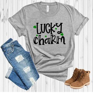 May include: A gray t-shirt with the text "LUCKY charm" in black with green shamrocks. The shirt is paired with blue jeans and brown boots.