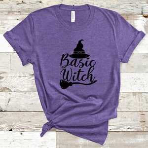 May include: A purple t-shirt with the words "Basic Witch" in a black script font, accompanied by a witch hat and broom graphic. The shirt is made of a soft material and is tied at the bottom.