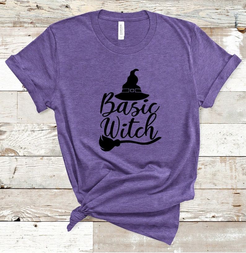 May include: A purple t-shirt with the words "Basic Witch" in a black script font, accompanied by a witch hat and broom graphic. The shirt is made of a soft material and is tied at the bottom.