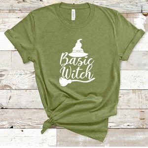 May include: Olive green t-shirt with the words "Basic Witch" in white script, accompanied by a witch hat and broom graphic. The shirt is made of a soft material and has rolled-up sleeves. The shirt is knotted at the bottom.