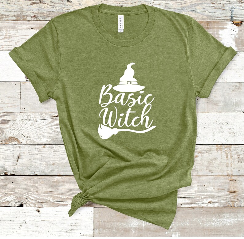 May include: Olive green t-shirt with the words "Basic Witch" in white script, accompanied by a witch hat and broom graphic. The shirt is made of a soft material and has rolled-up sleeves. The shirt is knotted at the bottom.