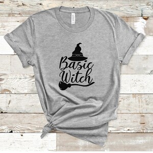 May include: A heather gray t-shirt with the words "Basic Witch" in black script, accompanied by a witch hat and broom graphic. The shirt is made of a soft material and has rolled-up sleeves.