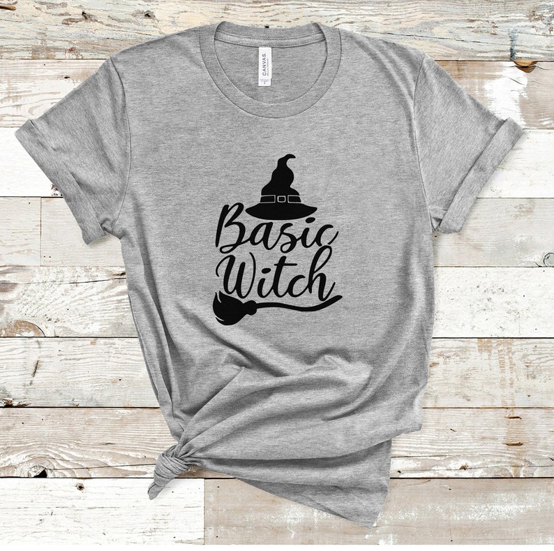 May include: A heather gray t-shirt with the words "Basic Witch" in black script, accompanied by a witch hat and broom graphic. The shirt is made of a soft material and has rolled-up sleeves.