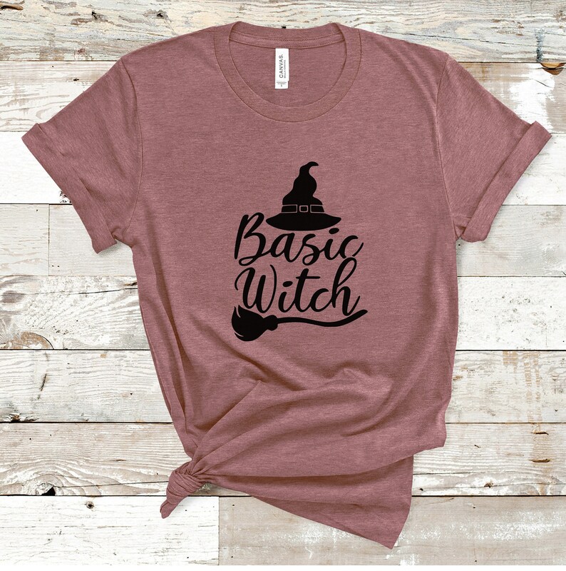 May include: A mauve-colored t-shirt with the words "Basic Witch" in black script, accompanied by a witch hat and broom graphic. The shirt is made of a soft material and has rolled-up sleeves.