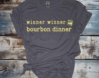 May include: A dark gray t-shirt with the text "winner winner bourbon dinner" in yellow lettering. A yellow illustration of a glass of bourbon is next to the text.