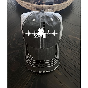 May include: Black and gray trucker hat with a white graphic of a horse and rider in a barrel racing design, with a heartbeat line. The hat has a distressed look with white stitching and a mesh back.
