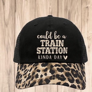 May include: Black trucker hat with a leopard print brim. The hat features the text "could be a TRAIN STATION KINDA DAY" in white script. The hat has a mesh back and a curved brim.