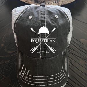 May include: Black and white distressed baseball cap with a white embroidered equestrian logo featuring crossed riding crops and a riding helmet. The text "EQUESTRIAN" is embroidered below the logo.