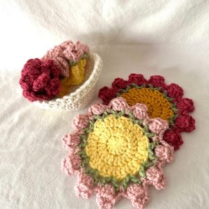 May include: Crochet flower coasters in a handmade design. The coasters have a yellow centre, with pink and burgundy petals, and green detailing. A small basket contains more coasters in matching colours. The items are shown on a white surface.