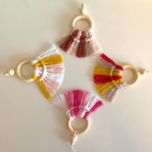 Puede incluir: Cuatro adornos de macramé con aros de madera y detalles de cuentas. Cada uno presenta una combinación de colores diferente: amarillo, rosa, blanco, rojo y marrón. Los adornos están dispuestos en forma de diamante sobre un fondo blanco.