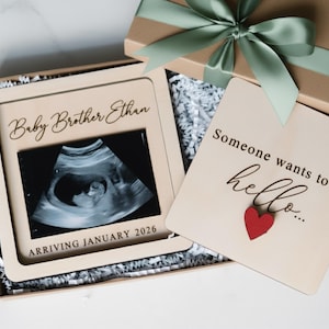 May include: A wooden picture frame with a sonogram image, the text "Baby Brother Ethan" and "ARRIVING JANUARY 2026". A gift box with a green ribbon and a wooden card that says "Someone wants to say hello" with a red heart.