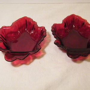 May include: Two ruby red glass leaf-shaped dishes. The dishes have a scalloped edge and a deep bowl. The glass is transparent, allowing light to pass through. These decorative dishes are ideal for serving small portions of food or as decorative accents.