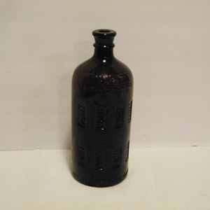 May include: A dark brown antique bottle with a cylindrical body and a narrow neck. The bottle has embossed rectangular panels around its sides and a slightly rounded base. The bottle is made of glass.
