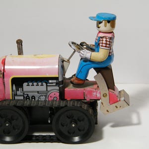 May include: Vintage toy tractor with a pink and yellow body, black treads, and a driver figure. The driver wears a blue cap, overalls, and a plaid shirt. The tractor has a detailed engine design and a metal exhaust pipe.