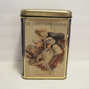May include: Rectangular tin with a gold lid and blue trim. The front features an illustration of three people in vintage clothing, with goggles and hats. Text on the tin reads "THE LADIES' HOME JOURNAL" and "AUGUST 1914".
