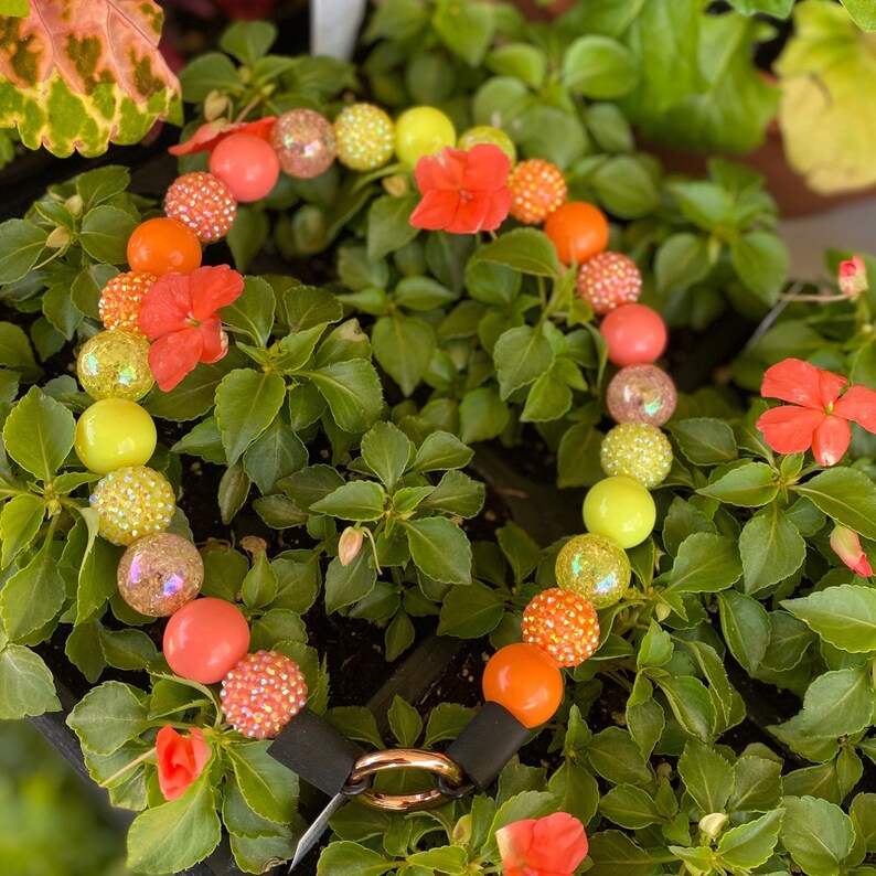 May include: A colorful beaded necklace with a mix of orange, yellow, and pink beads. The necklace is made of large, round beads with a glossy finish. The necklace is lying on a bed of green leaves.