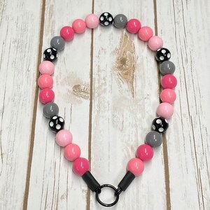 May include: A pink and gray beaded necklace with black polka dots. The necklace has a metal ring clasp.