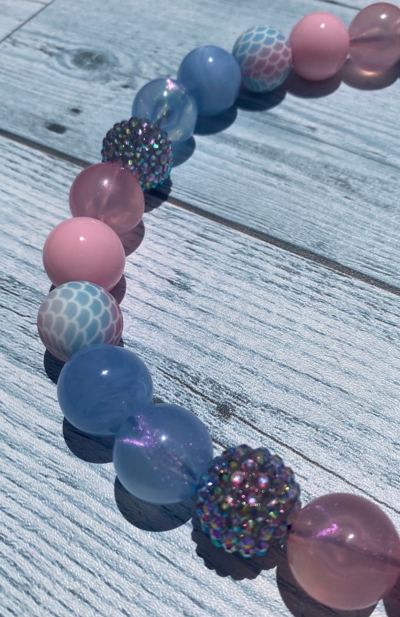 May include: A colorful beaded necklace with pink, blue, and iridescent beads. The beads are arranged in a pattern and have a variety of textures.