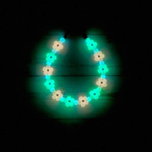 May include: A necklace with flower-shaped beads that glow. The beads alternate between white and bright green, forming a circular design. The necklace is set against a dark background, emphasizing the vibrant colours of the beads.