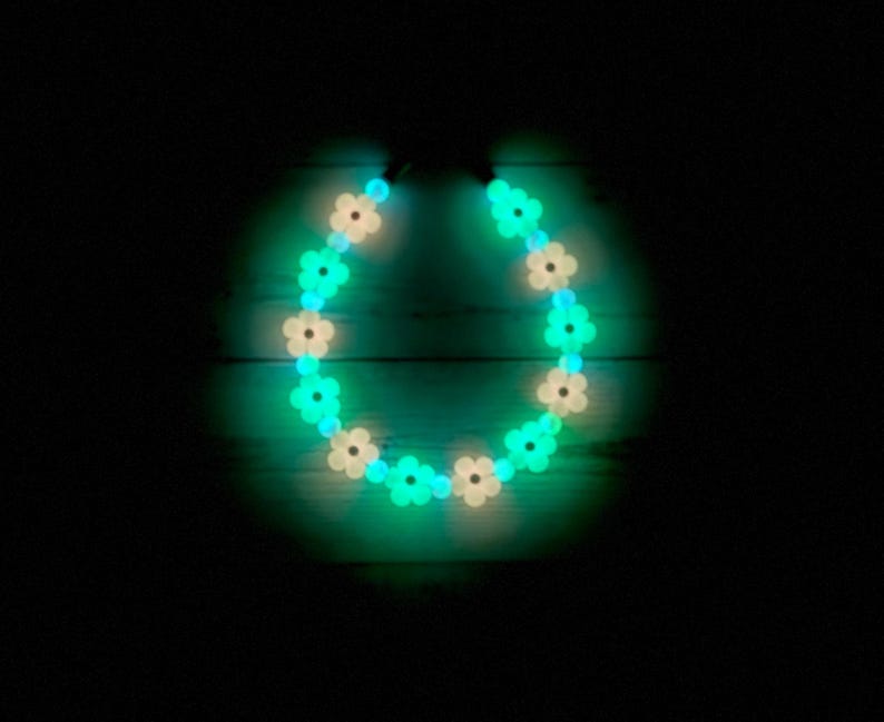 May include: A necklace with flower-shaped beads that glow. The beads alternate between white and bright green, forming a circular design. The necklace is set against a dark background, emphasizing the vibrant colours of the beads.