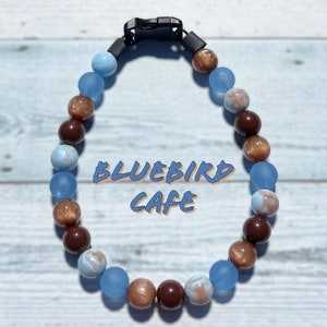 May include: A blue and brown beaded necklace with a black clasp. The beads are made of glass and have a frosted finish. The necklace is on a white wooden background. The text "BLUEBIRD CAFE" is visible in the center of the image.