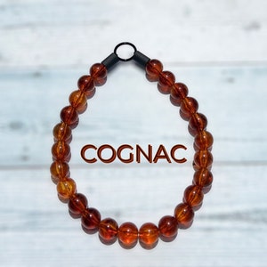 Puede incluir: Un collar de cuentas de ámbar color coñac con un cierre de metal negro. El collar está sobre un fondo de madera blanca.