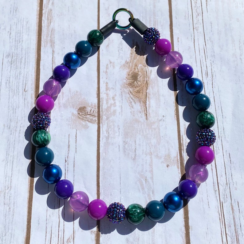 May include: A colorful beaded necklace with shades of purple, pink, blue, and green. The beads are round and have a glossy finish.