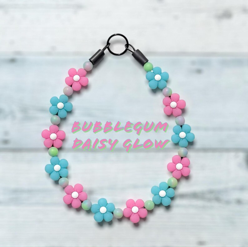 May include: A necklace featuring pink and blue daisy flower beads, with light green and white beads. The necklace has a black clasp and the text "BUBBLEGUM DAISY GLOW" in the center.