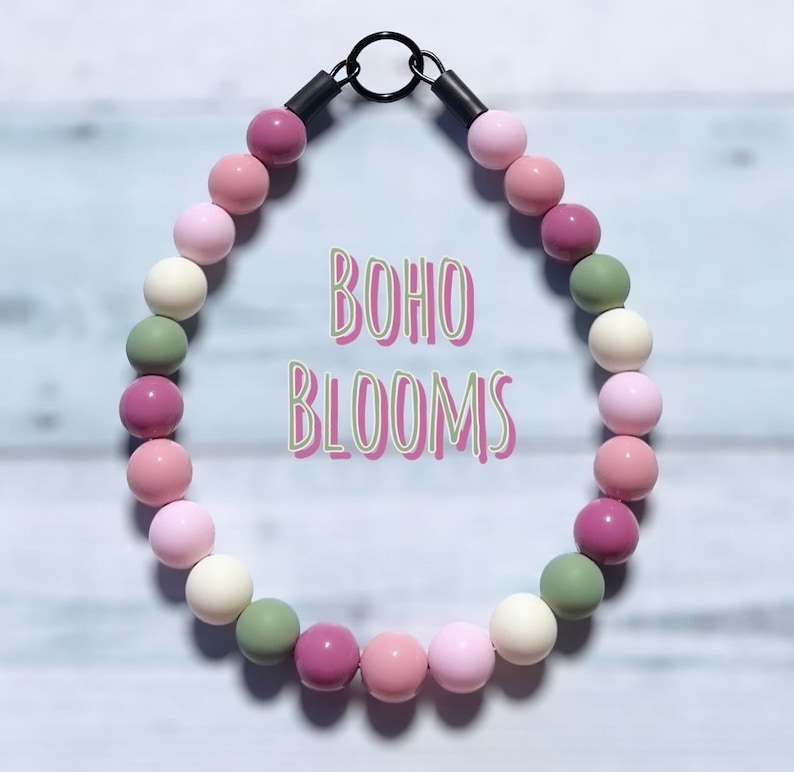 May include: A beaded necklace with a variety of pastel-colored beads, including pink, white, green, and mauve. The necklace has a black metal clasp and the words "BOHO BLOOMS" in a whimsical font.