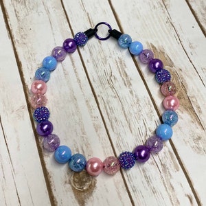 May include: A colorful beaded necklace with a gradient of pink, blue, and purple beads. The beads are made of various materials, including glass, plastic, and metal. The necklace has a metal clasp.