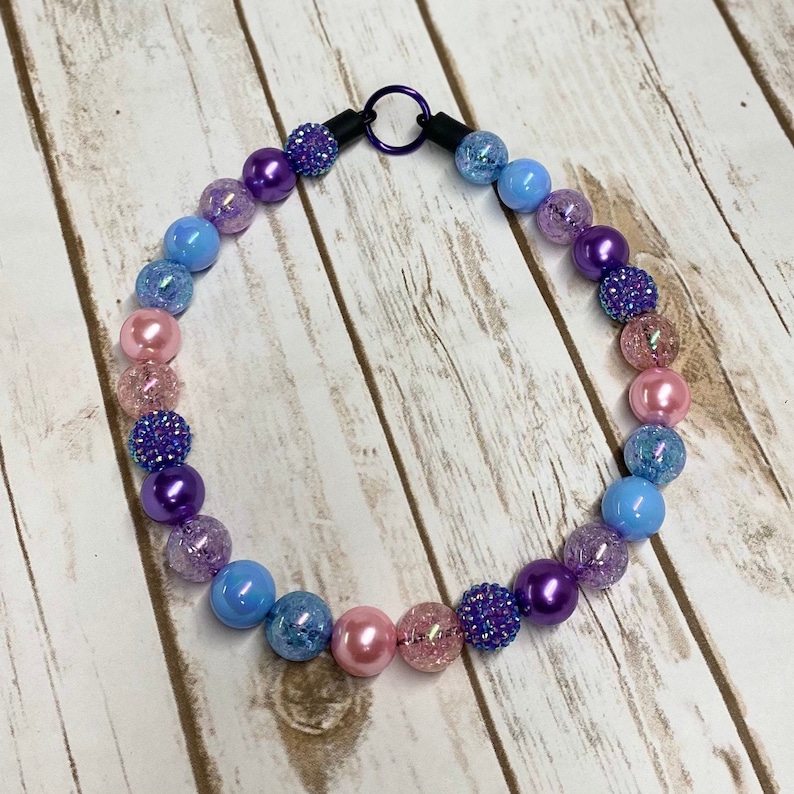 May include: A colorful beaded necklace with a gradient of pink, blue, and purple beads. The beads are made of various materials, including glass, plastic, and metal. The necklace has a metal clasp.