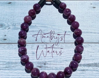 May include: A purple amethyst gemstone beaded necklace with a rose gold clasp. The necklace is on a white wooden background.