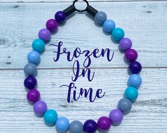 Puede incluir: Un collar de cuentas de colores con un cierre negro. Las cuentas son de color morado, azul y gris. El collar tiene el texto "Frozen in Time" escrito en él.