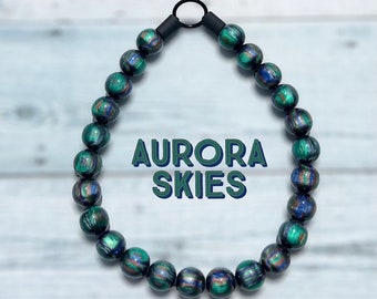May include: A necklace made of green, blue, and black beads with a black clasp. The beads have a swirling pattern that resembles an aurora borealis. The text "AURORA SKIES" is printed in the center of the image.