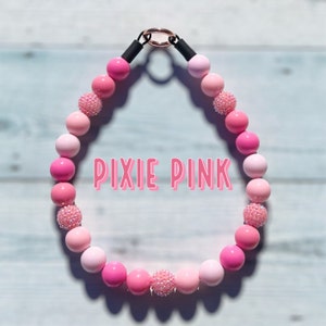 Puede incluir: Un collar de perro de cuentas rosadas con un cierre negro y un dije plateado. Las cuentas son de varios tonos de rosa y algunas tienen brillo. El collar está etiquetado como "PIXIE PINK".