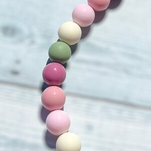 May include: A necklace featuring a series of colorful, round beads in shades of pink, green, and cream. The beads are strung together, creating a playful and eye-catching accessory. The necklace is shown against a light blue wooden background.