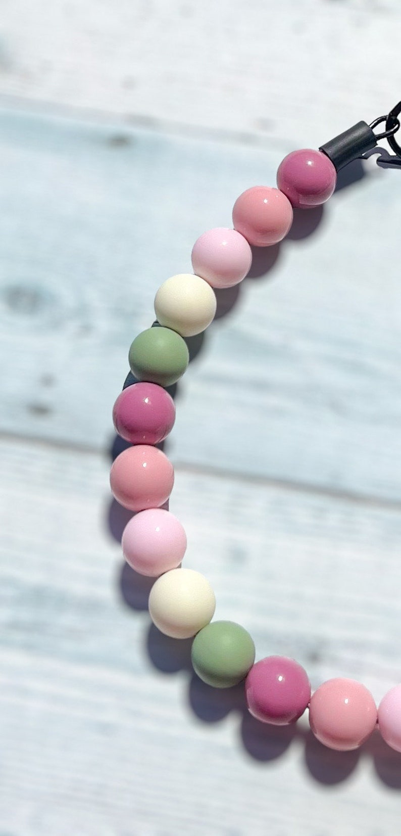 May include: A necklace featuring a series of colorful, round beads in shades of pink, green, and cream. The beads are strung together, creating a playful and eye-catching accessory. The necklace is shown against a light blue wooden background.
