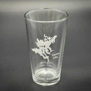 May include: Clear glass pint glass with a detailed etched map of Squam Lake. The glass is cylindrical with a slightly flared top. The etched design is in white and the text "Squam Lake" is below the map.