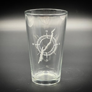 May include: A clear pint glass with a compass rose design and a map of a lake. The compass rose features the cardinal directions, and the lake map is etched in white. The glass is empty and sits on a dark surface.