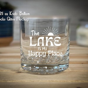 May include: A clear rocks glass with the text "The Lake is my Happy Place" in white. The glass has a knob bottom and is approximately 10.25 oz. The glass is on a wooden surface.