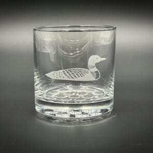 May include: A clear, round drinking glass with a detailed etched image of a loon. The loon is depicted in white, with a textured body and a long neck. The glass has a thick base.