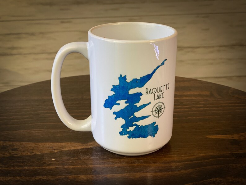 May include: White ceramic coffee mug with a blue watercolor illustration of the outline of a lake and the text "Raquette Lake".