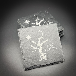 May include: Two square slate coasters with a map of Lake Burton engraved in white. Each coaster features a compass rose and the lake's name. The coasters are dark gray with a textured surface.