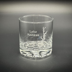 May include: A clear glass tumbler with a textured bottom. The glass is engraved with the words "Lake Keowee" and an outline of the lake.