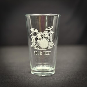 May include: Clear glass pint glass with a detailed etched design of a drum set. The text "YOUR TEXT" is etched below the drum set image. The glass is empty and sits on a dark surface.