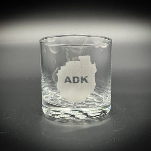 May include: A clear glass tumbler with a raised bottom and an etched design of the Adirondack Mountains with the text "ADK" inside the outline of the mountains.