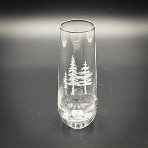 May include: A clear, tall glass with a forest tree design etched on the front. The glass is cylindrical with a rounded base and a smooth rim. The etched design features three stylized pine trees in white.