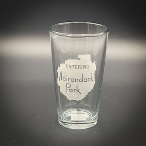 May include: A clear glass pint glass with an etched design of the Adirondack Park outline and the text "Entering Adirondack Park".