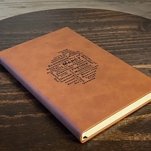 May include: A brown leather journal with a diamond-shaped design of mountain names in black text. The journal is closed and has a cream-colored edge. The cover has a textured appearance and is set on a wooden surface.