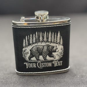 May include: Black leather flask with a silver top and a laser engraved design of a bear walking through a forest. The text "Your Custom Text" is engraved below the bear.